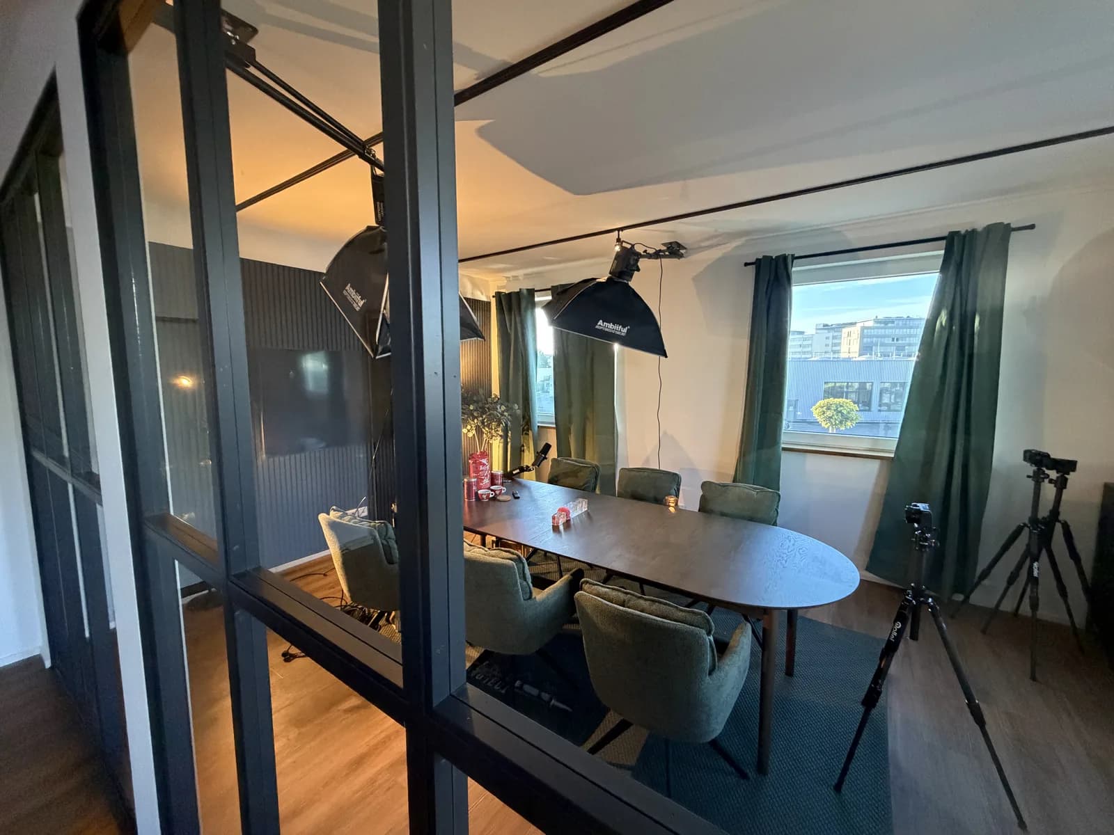Blick durch die schwarz gerahmte Glaswand in den Aufnahmeraum, im Raum ein Kamerastativ, Softboxen und Fenster mit Stadtblick