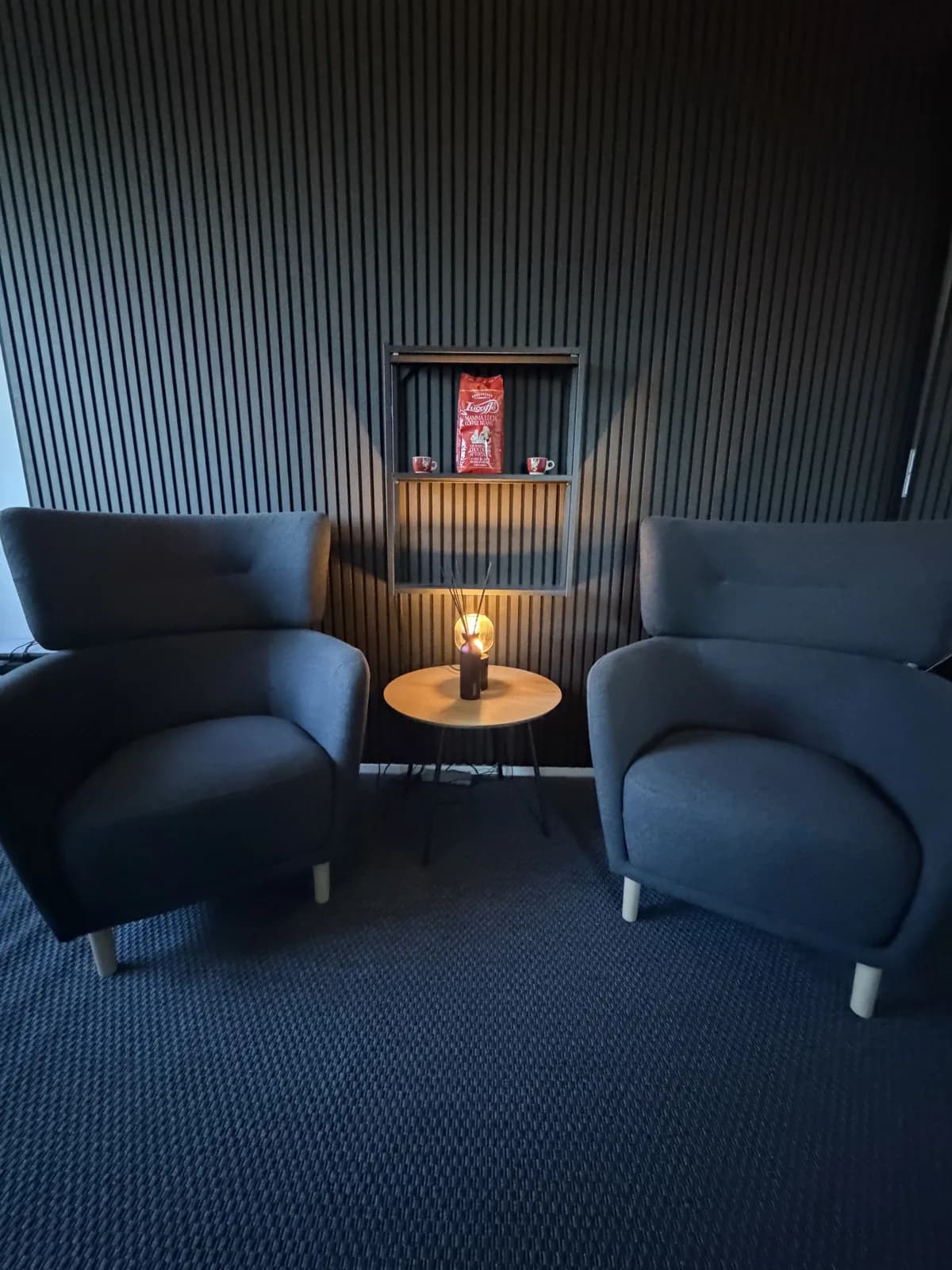 Lounge-Ecke mit zwei dunkelblauen Sesseln, kleinem runden Holztisch, warmer Glühlampe und Regalnische an einer Lamellenwand
