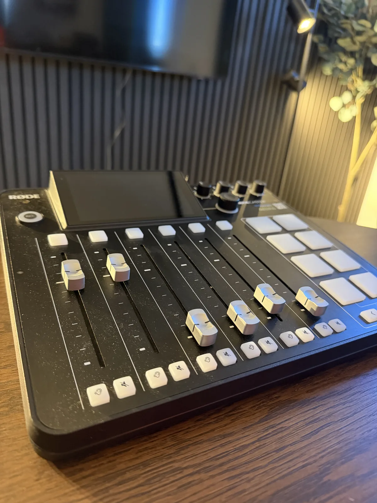 RØDE RODECaster Pro II Podcast-Mischpult mit Fadern, Touchscreen und Pads auf dem Holztisch, dahinter die Lamellenwand